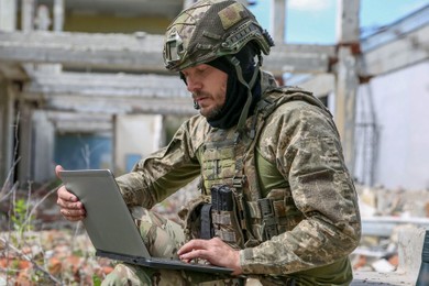 Military mission. Soldier in uniform using laptop near abandoned building outside