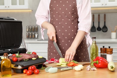 Woman cooking different products with electric grill at wooden table in kitchen, closeup