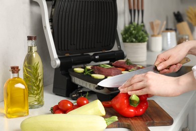 Woman cooking different products with electric grill at white wooden table in kitchen, closeup