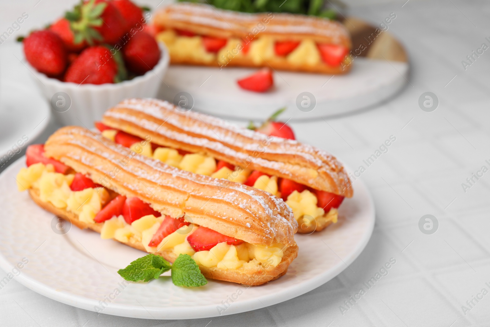 Photo of Delicious eclairs filled with cream, strawberries and mint on white tiled table, closeup