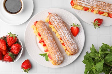 Photo of Delicious eclairs filled with cream, strawberries, mint and tea on white tiled table, flat lay
