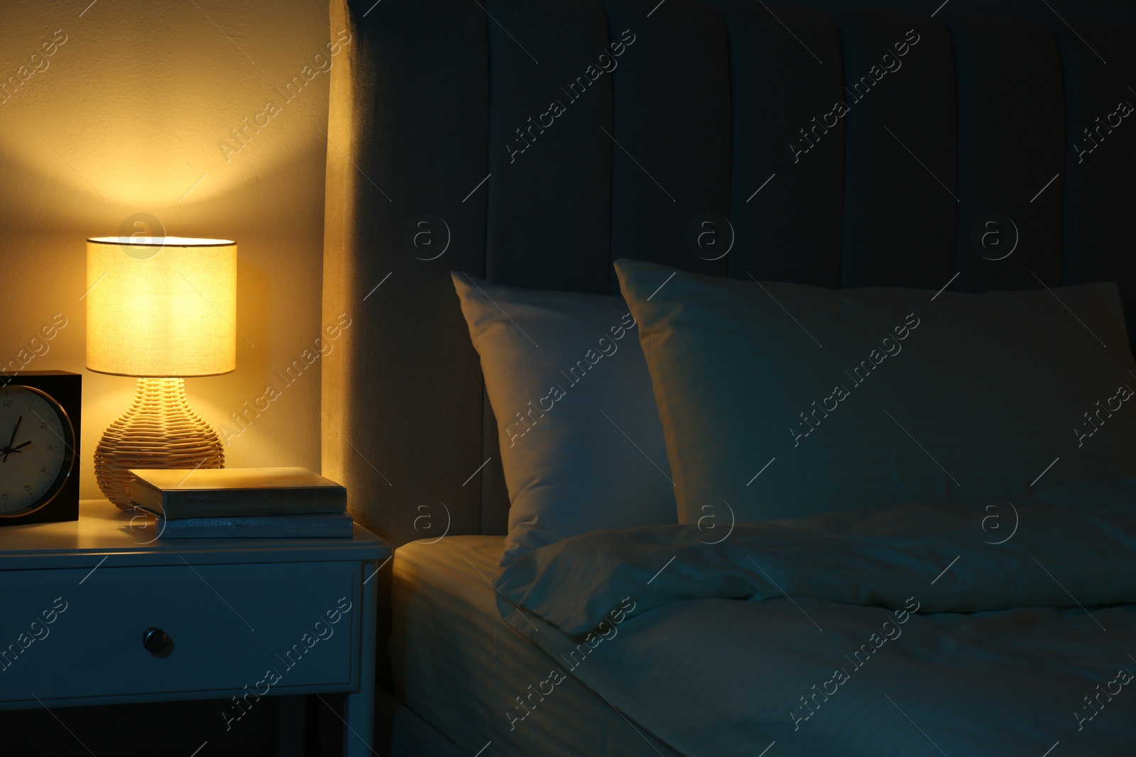 Photo of Nightlight, clock and books on bedside table near bed indoors