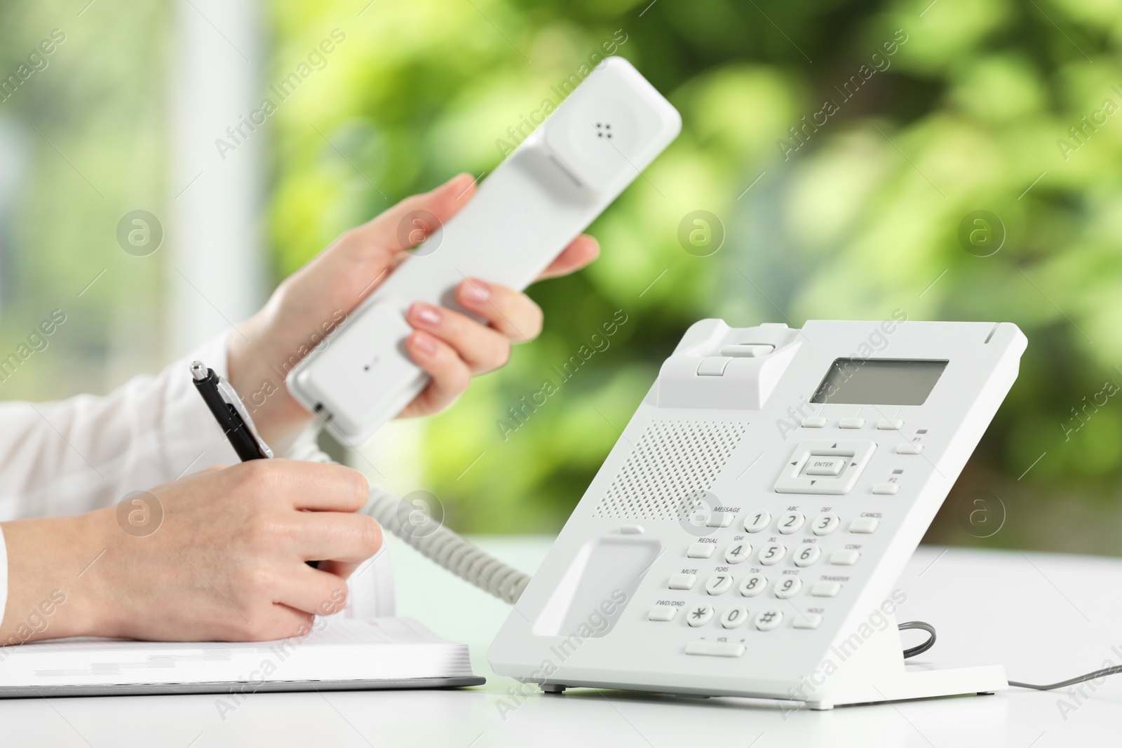 Photo of Assistant with telephone handset writing at white table against blurred green background, closeup