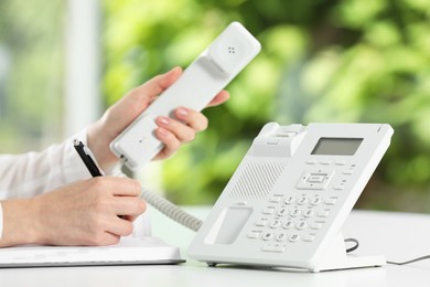 Assistant with telephone handset writing at white table against blurred green background, closeup