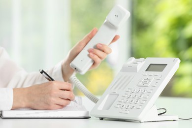 Assistant with telephone handset writing at white table against blurred green background, closeup