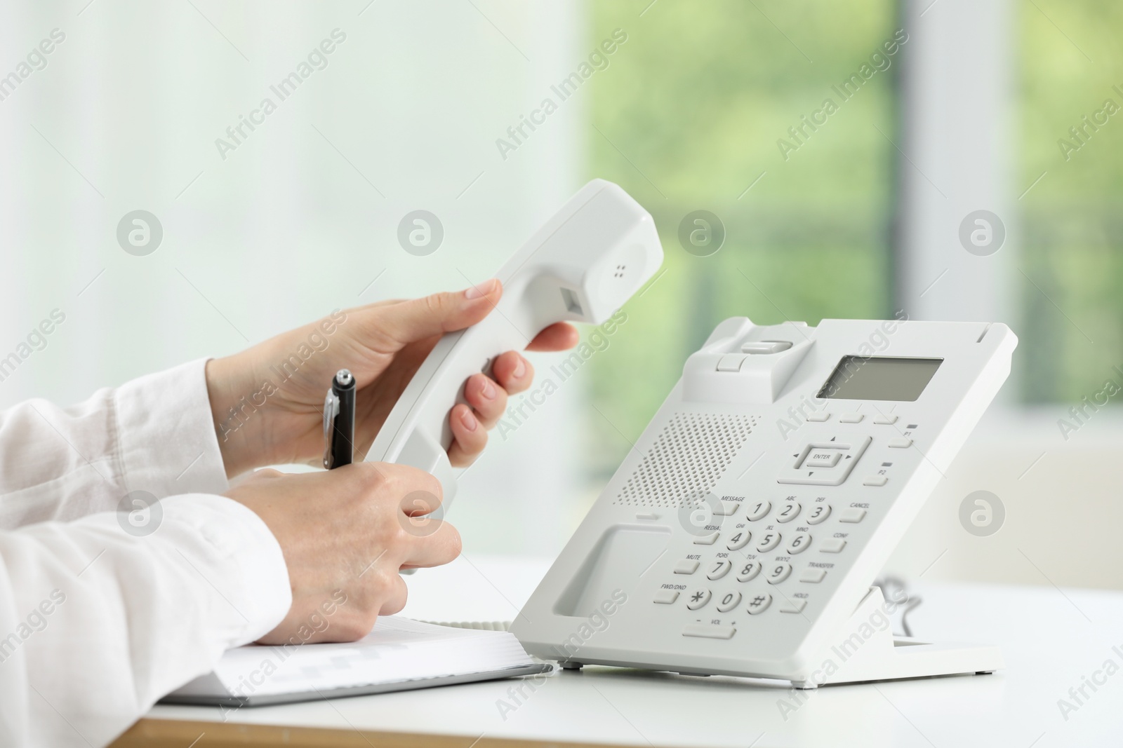 Photo of Assistant with telephone handset writing at white table against blurred background, closeup