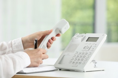 Assistant with telephone handset writing at white table against blurred background, closeup