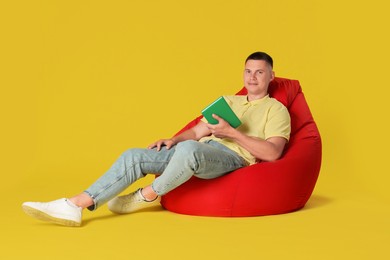 Handsome man with book on red bean bag chair against yellow background