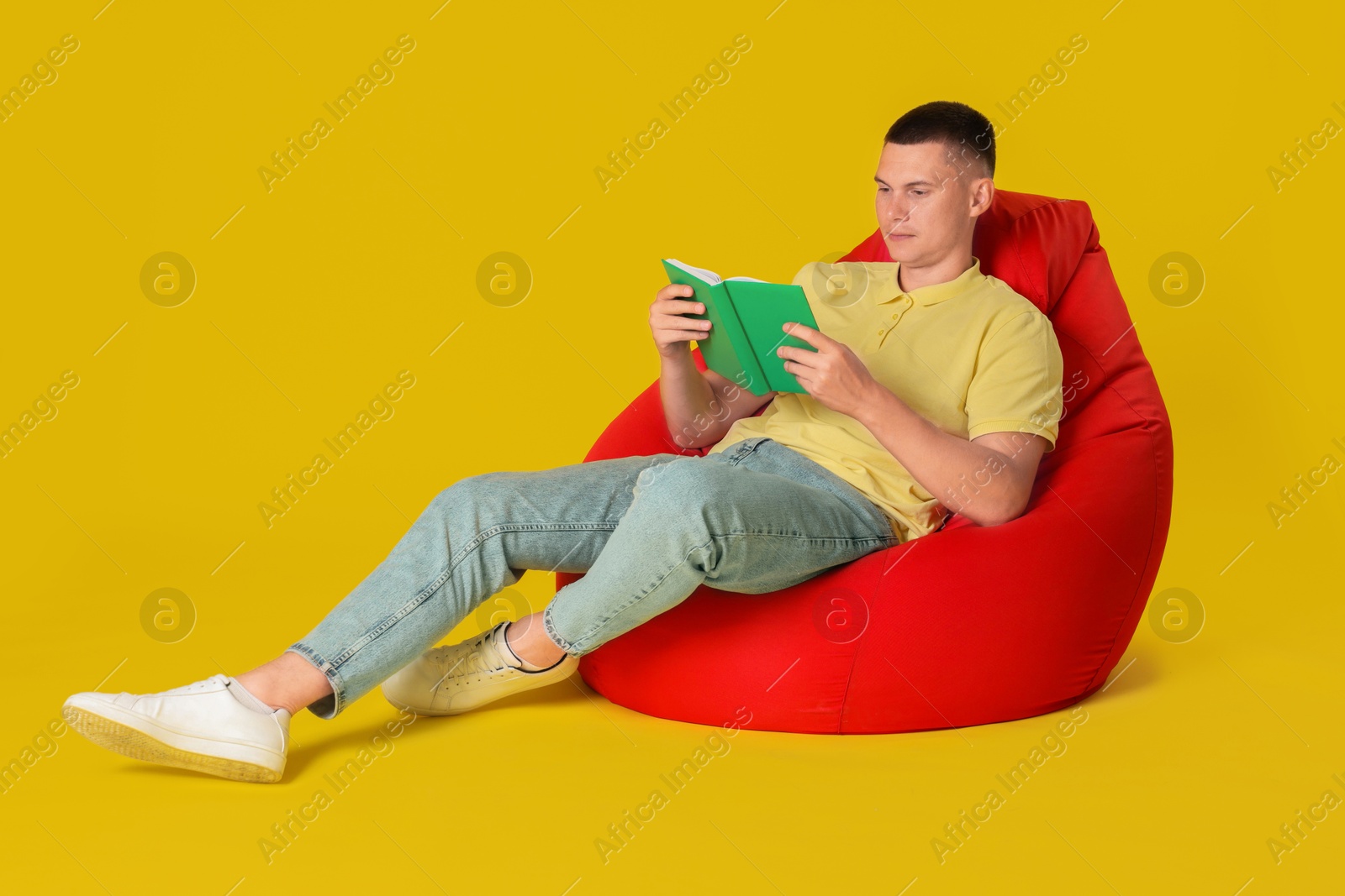 Photo of Handsome man reading book on red bean bag chair against yellow background