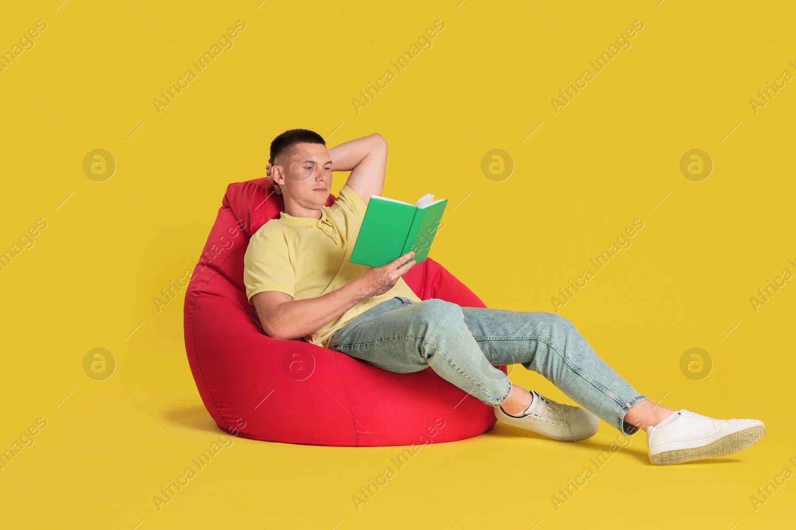 Photo of Handsome man reading book on red bean bag chair against yellow background