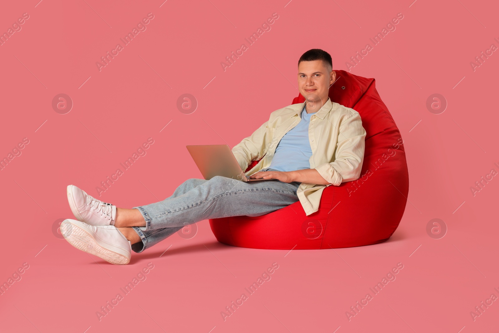 Photo of Handsome man with laptop on red bean bag chair against pink background