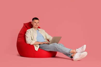 Handsome man with laptop on red bean bag chair against pink background