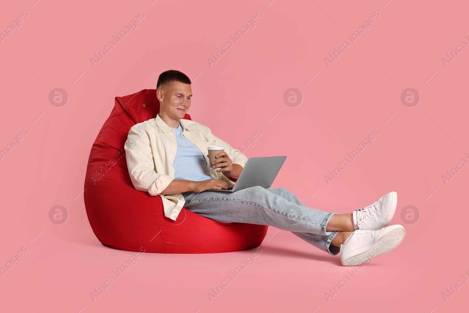 Photo of Handsome man with laptop and paper cup on red bean bag chair against pink background