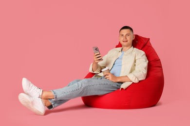 Handsome man with smartphone and paper cup on red bean bag chair against pink background