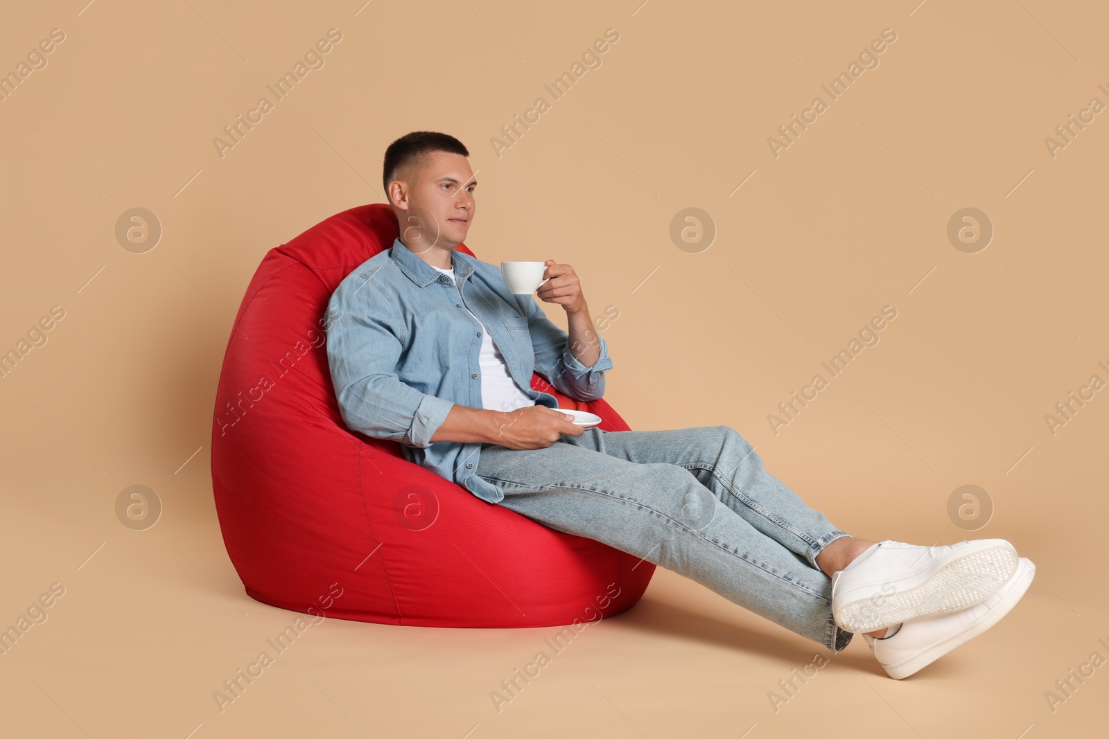 Photo of Handsome man drinking coffee on red bean bag chair against beige background