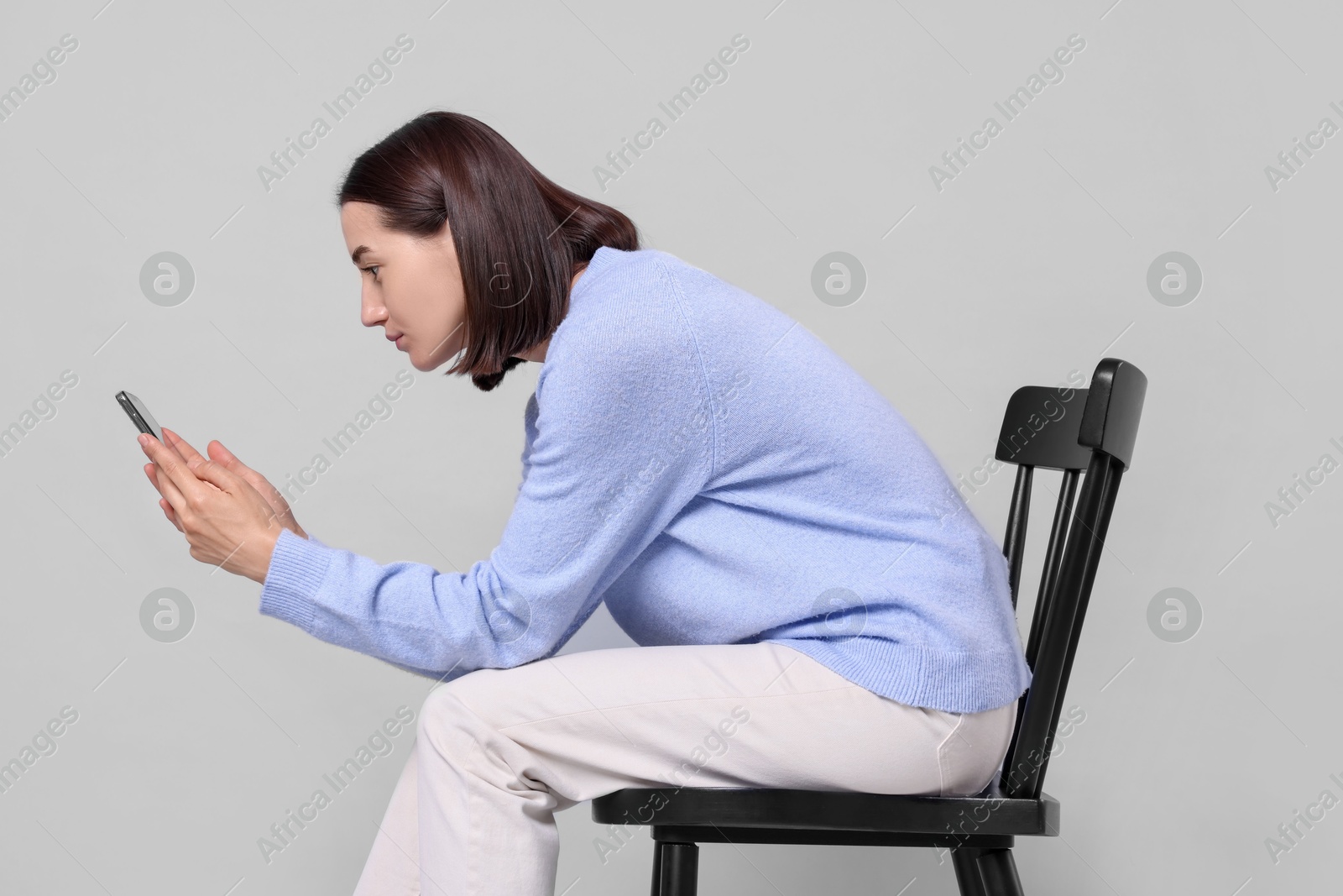 Photo of Woman with poor posture sitting on chair and using smartphone against gray background