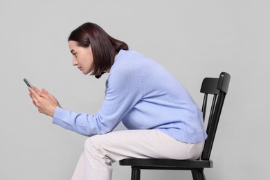 Woman with poor posture sitting on chair and using smartphone against gray background