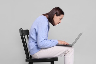 Woman with poor posture sitting on chair and using laptop against gray background