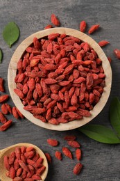 Photo of Dried goji berries in bowl, spoon and leaves on grey textured table, flat lay