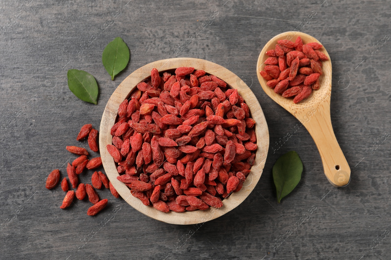 Photo of Dried goji berries in bowl, spoon and leaves on grey textured table, flat lay