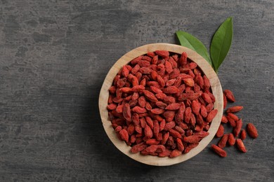 Photo of Dried goji berries in bowl and leaves on grey textured table, flat lay. Space for text