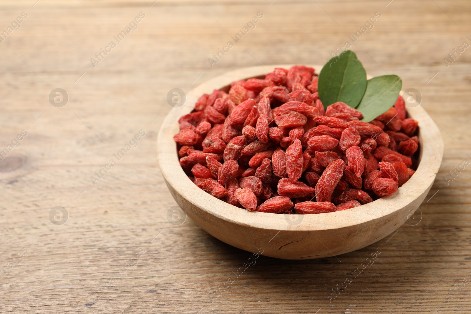 Photo of Dried goji berries and leaves in bowl on wooden table, closeup. Space for text