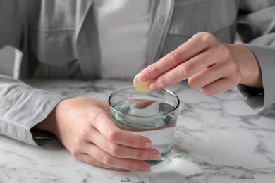 Woman putting effervescent pill into glass of water at white marble table, closeup