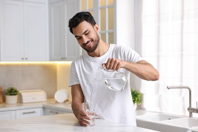 Morning of happy man pouring water from jug into glass at table in kitchen