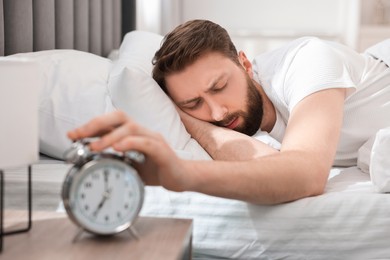 Photo of Sleepy young man turning off alarm clock in bedroom at morning