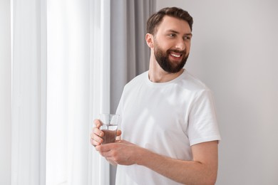 Photo of Happy young man with glass of water near window at morning