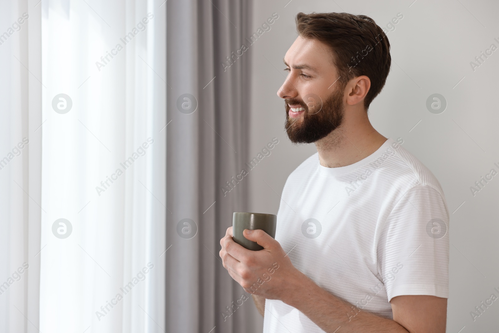 Photo of Happy young man with cup of coffee near window at morning