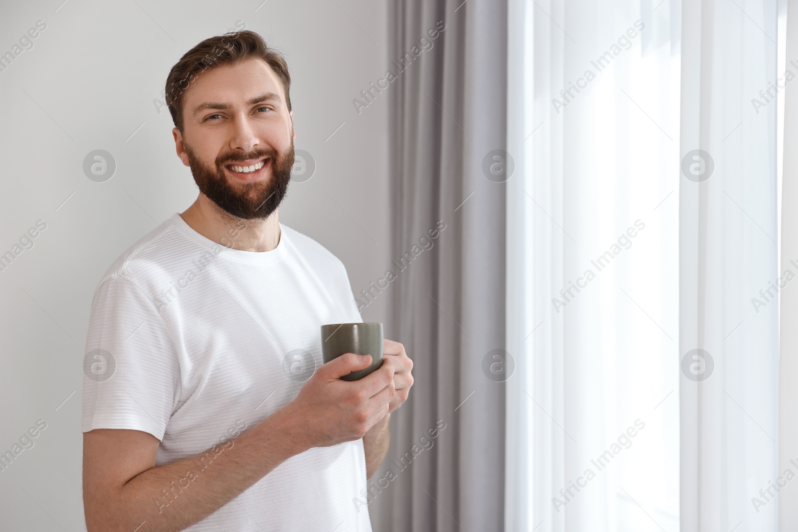 Photo of Happy young man with cup of coffee near window at morning