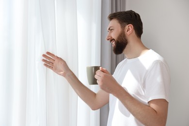 Photo of Happy young man with cup of coffee near window at morning