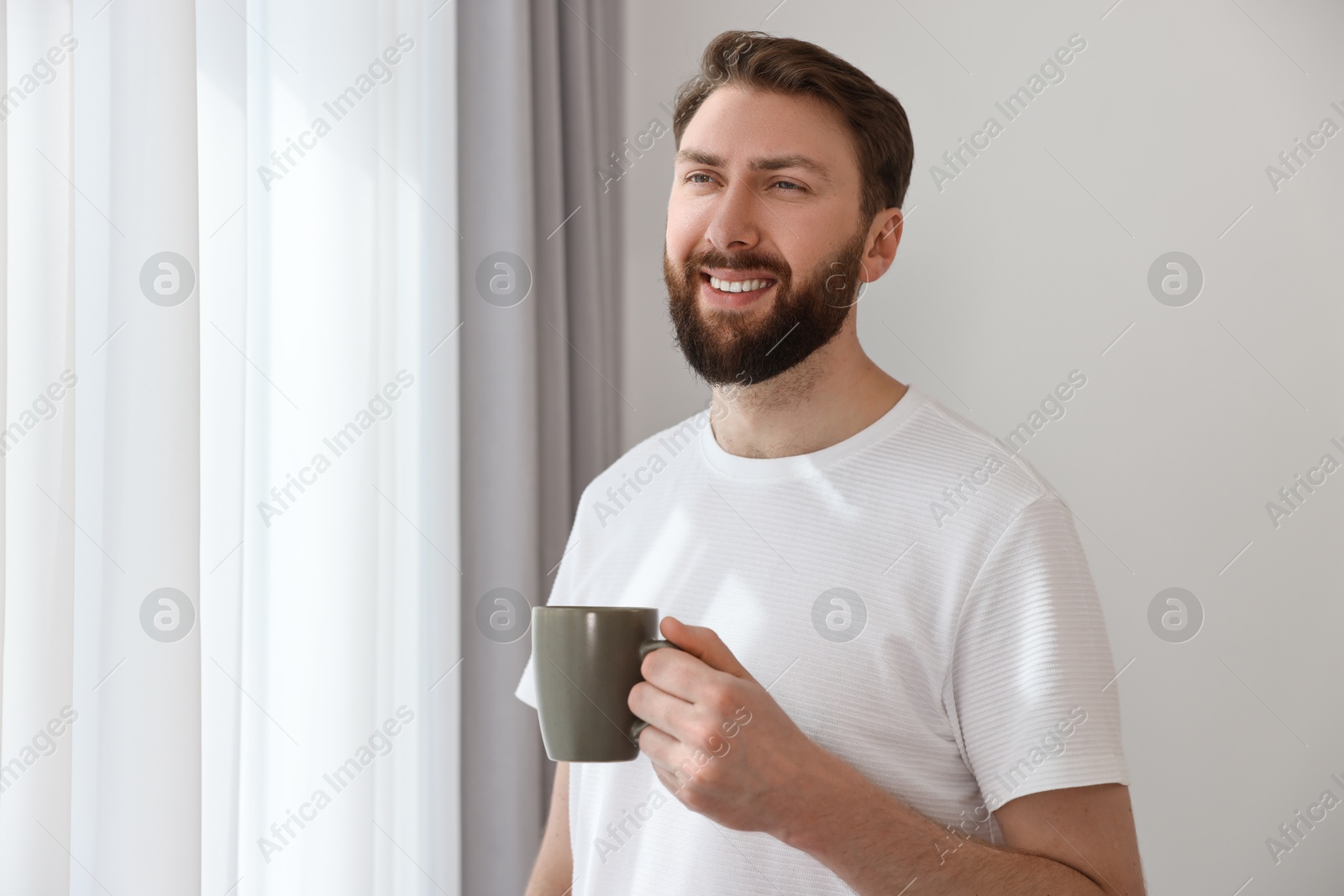 Photo of Happy young man with cup of coffee near window at morning