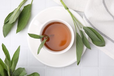 Aromatic herbal tea in cup with sage on white tiled table, flat lay