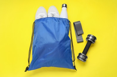 Blue drawstring bag, thermo bottle, sneakers, fitness elastic band and dumbbells on yellow background, flat lay