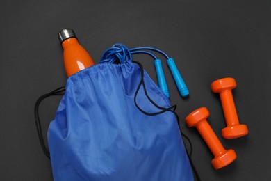 Photo of Blue drawstring bag, thermo bottle, skipping rope and dumbbells on black background, flat lay