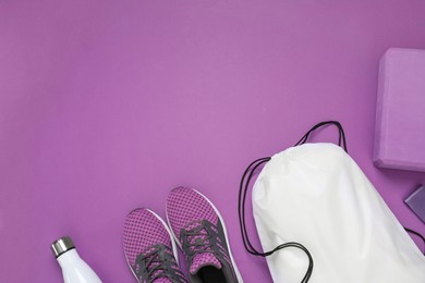 Photo of White drawstring bag, thermo bottle, sneakers and yoga block on purple background, flat lay. Space for text