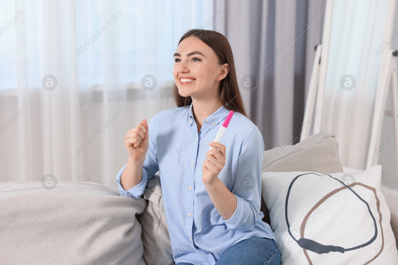 Photo of Happy woman holding pregnancy test on sofa indoors