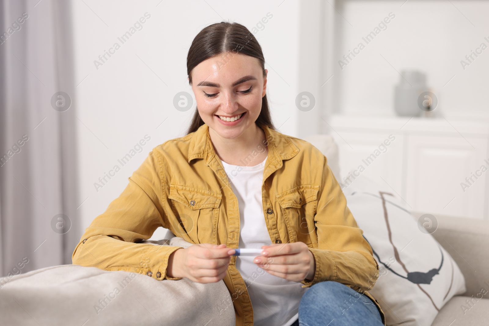 Photo of Happy woman holding pregnancy test on sofa indoors
