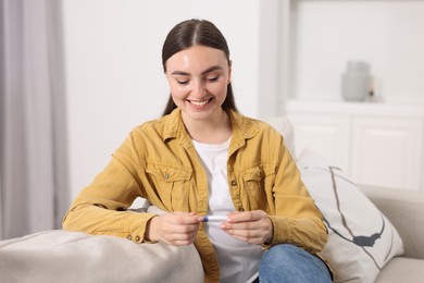Happy woman holding pregnancy test on sofa indoors