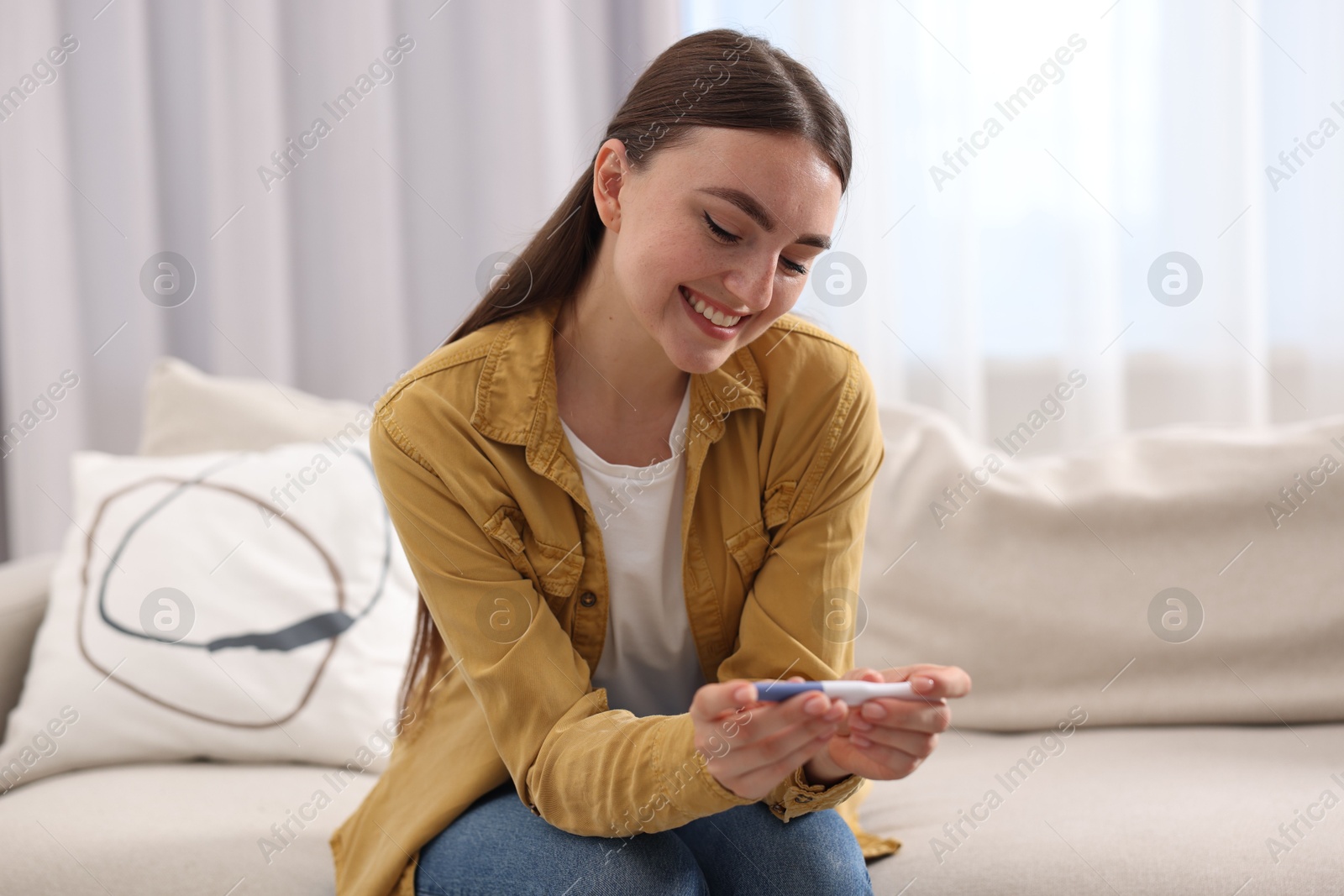 Photo of Happy woman holding pregnancy test on sofa indoors