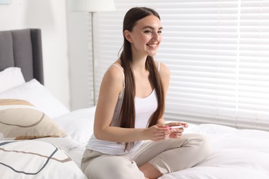 Happy woman holding pregnancy test on bed in room