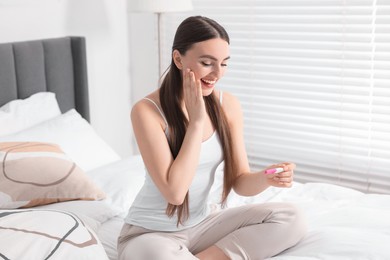 Happy woman holding pregnancy test on bed in room