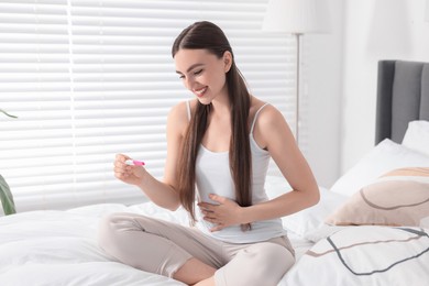 Happy woman holding pregnancy test on bed in room