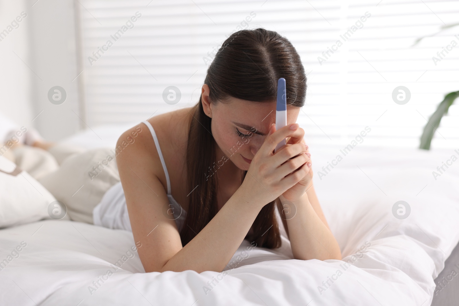 Photo of Sad woman holding pregnancy test on bed in room