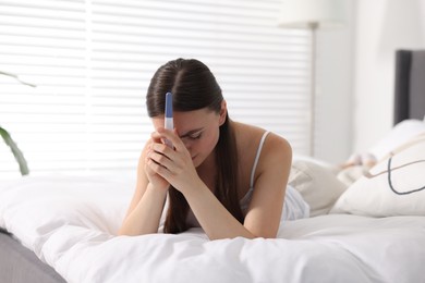 Sad woman holding pregnancy test on bed in room