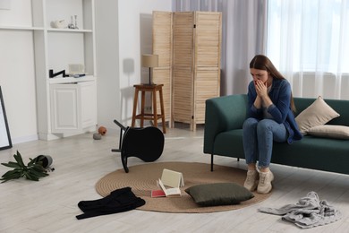 Photo of Desperate woman sitting on sofa in messy living room after robbery