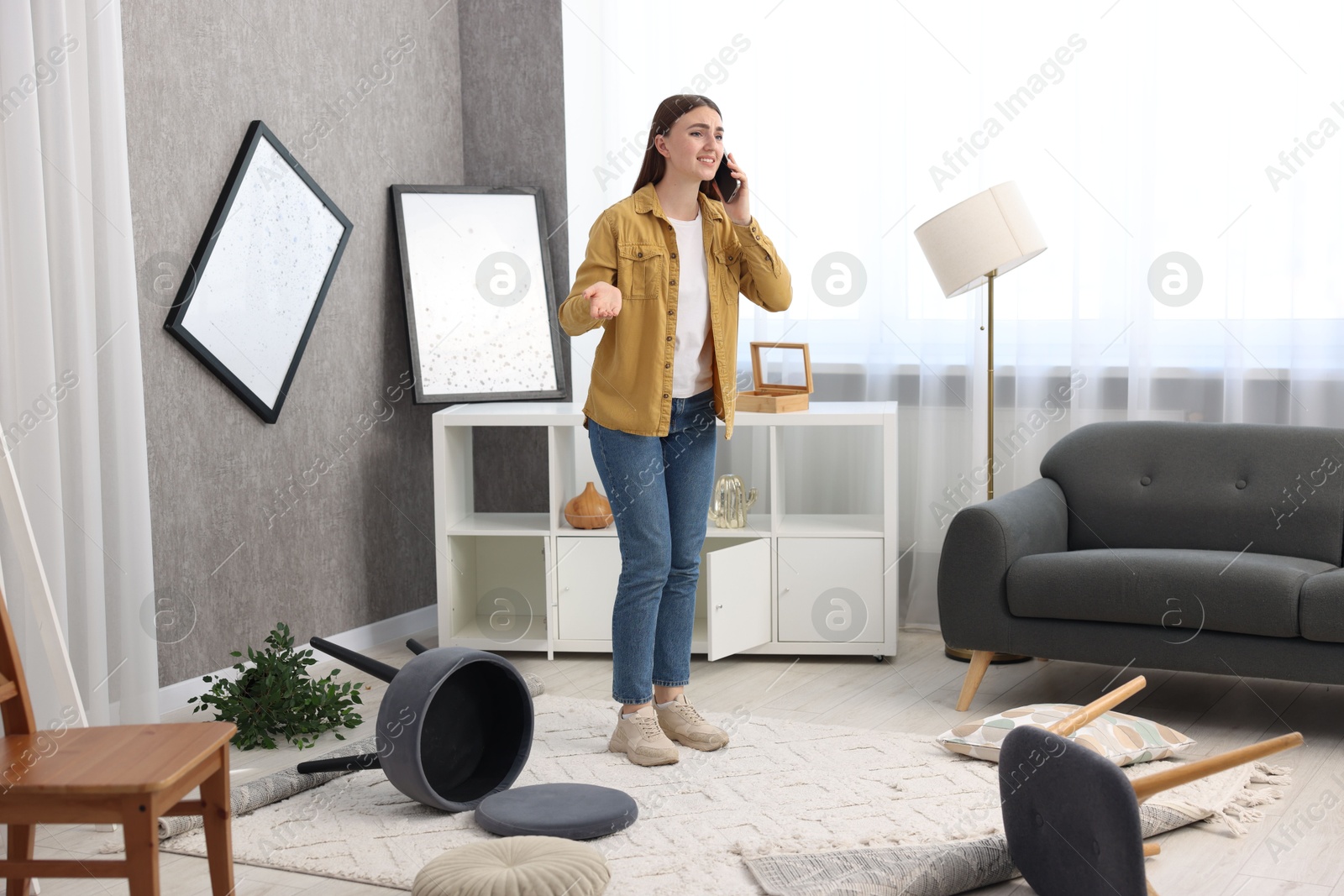 Photo of Desperate woman talking on smartphone in messy living room after robbery