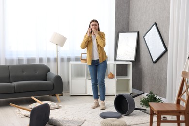 Photo of Desperate woman talking on smartphone in messy living room after robbery